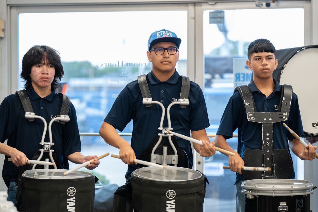 Three people play drums. One wears a cap and glasses, another has long hair, and the third is in the middle.