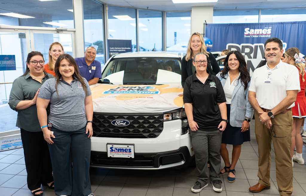 Nine people stand behind a decorated white Ford Explorer. A banner with a logo is visible behind them.