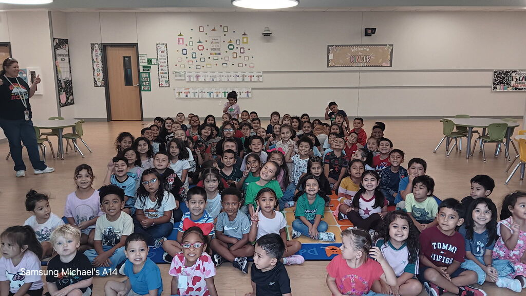 A large group of children sits on the floor in a classroom, with some standing in the background.
