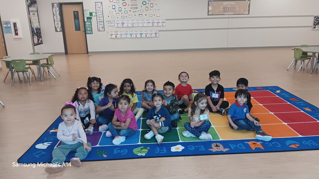 Group of children sitting on a colorful mat in a classroom. Tables and chairs are in the background.