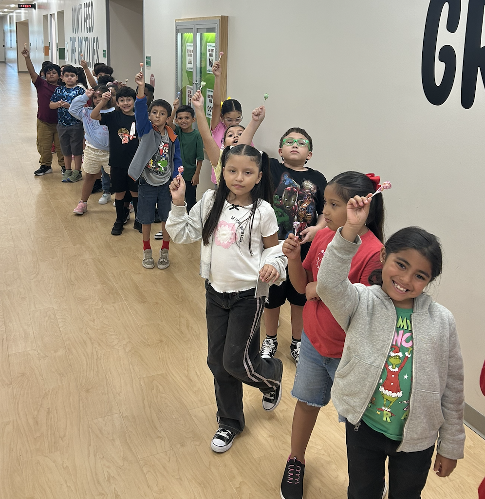 Children line up in a hallway, raising their hands. Some have toothbrushes. The wall says "GR".
