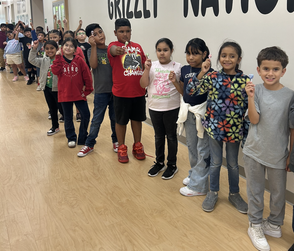 Nine children stand in a row in a hallway, smiling and holding toothbrushes, with a white wall reading "GRILLZI NATION" in the background.