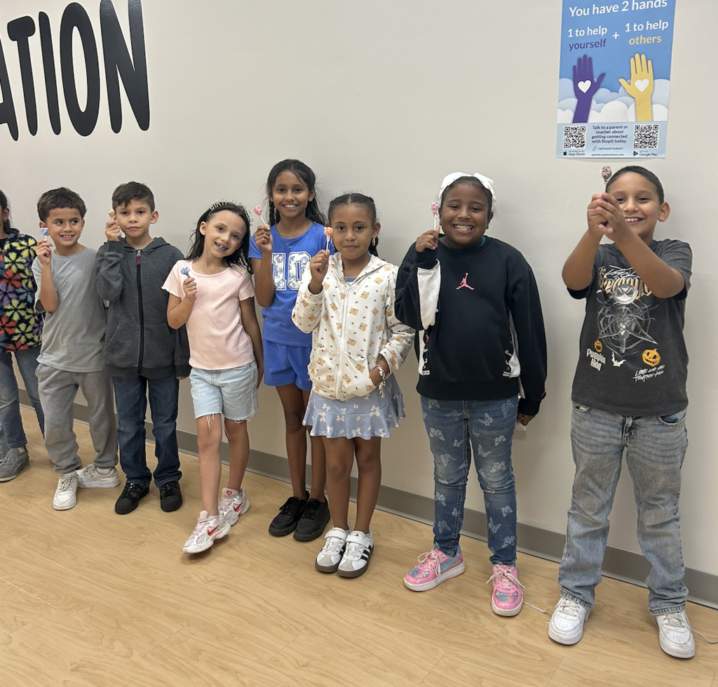 A group of children pose for a photo, standing in a line with a wall in the background.