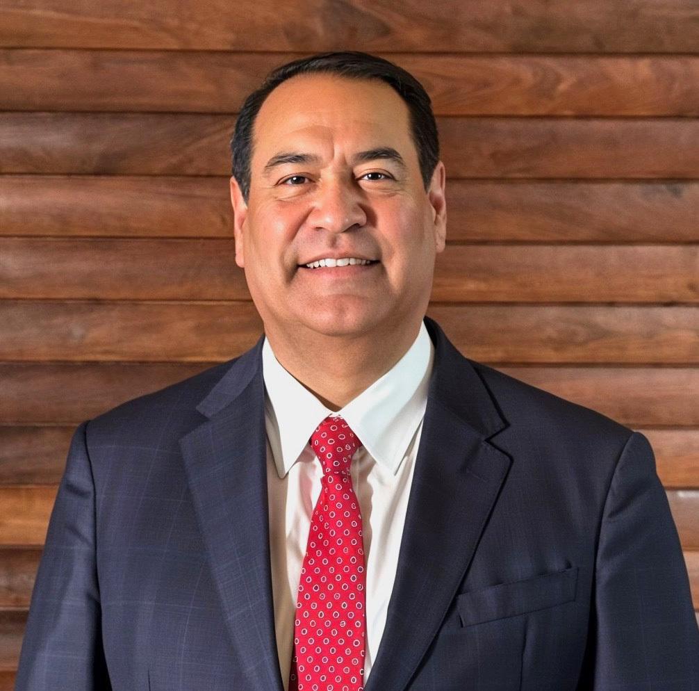 A man in a navy blue suit and red tie smiles in front of a wooden wall.