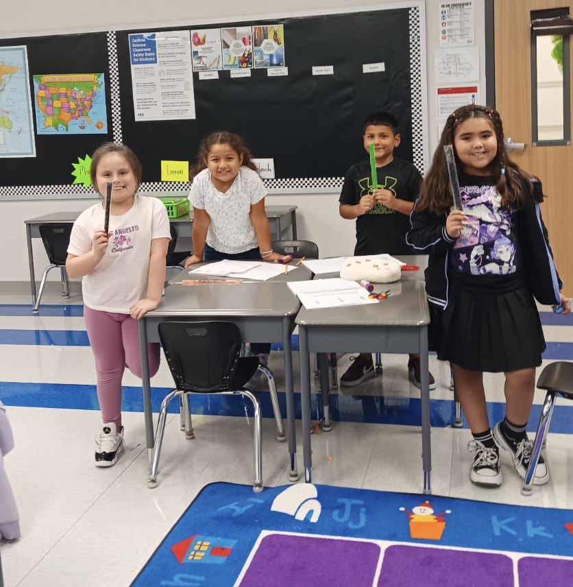 Four children in a classroom, holding items and smiling, standing by a desk with papers and a cake.