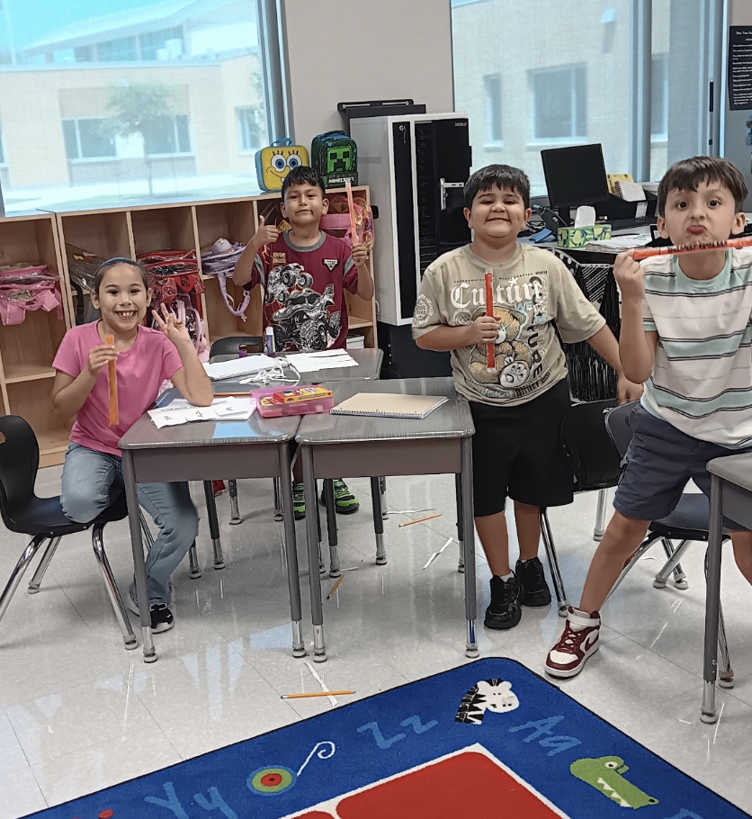 Four children in a classroom, three boys and one girl, seated around a table, smiling and holding up sticks.