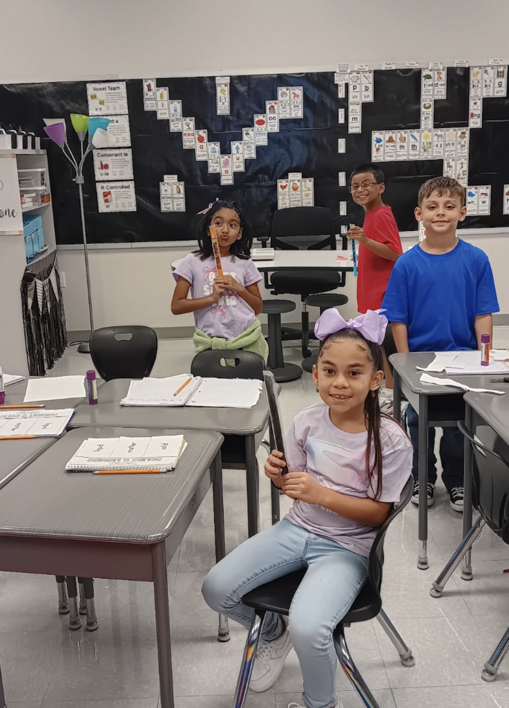 Four children in a classroom; one sits at a desk, another holds a recorder. Behind them, a blackboard displays various numbers.