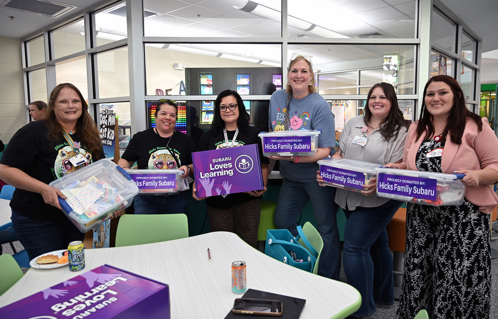 Five women in a meeting room holding purple boxes. Two more purple boxes are on the table in front of them.