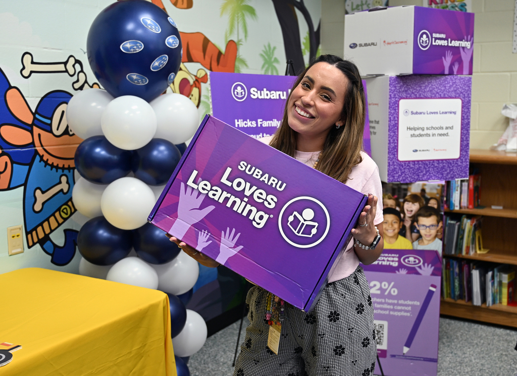 A woman holding a purple box labeled "Subaru Loves Learning" stands in a classroom with balloons.