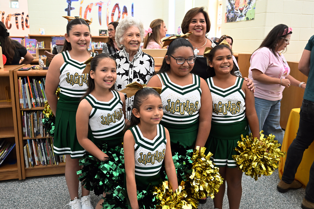 Five cheerleaders in green and white uniforms with gold pom-poms pose with an older woman. Behind them is a bookshelf.