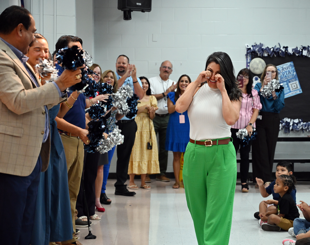 A woman in green pants walks among a group of adults and children in a room.