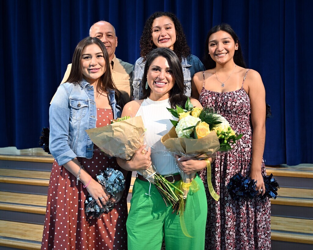 Five people on stairs; one holds flowers, smiling. A blue curtain in the background.