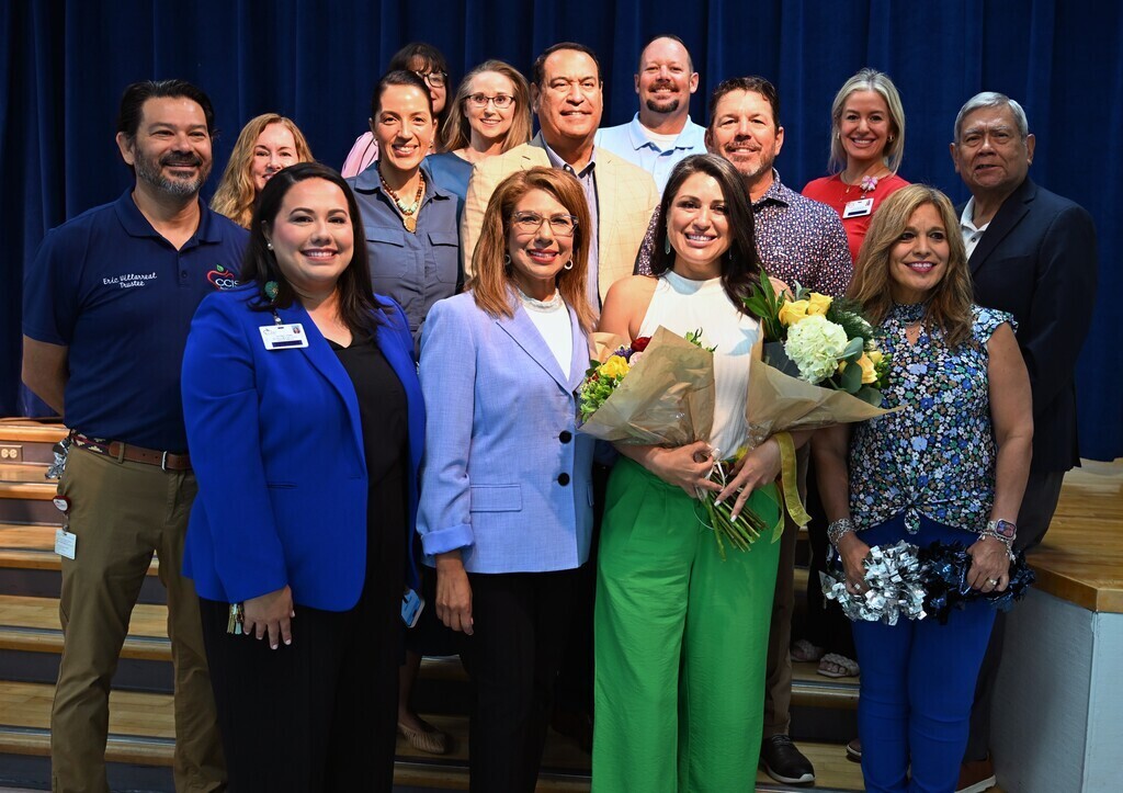 A group of adults, including women and men, stand together with one holding flowers, in a room with stairs and a blue curtain.