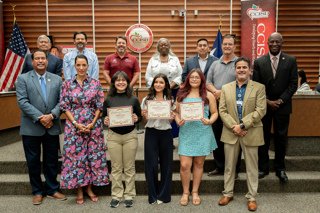 A group of people standing on a stage, holding certificates, with flags in the background.