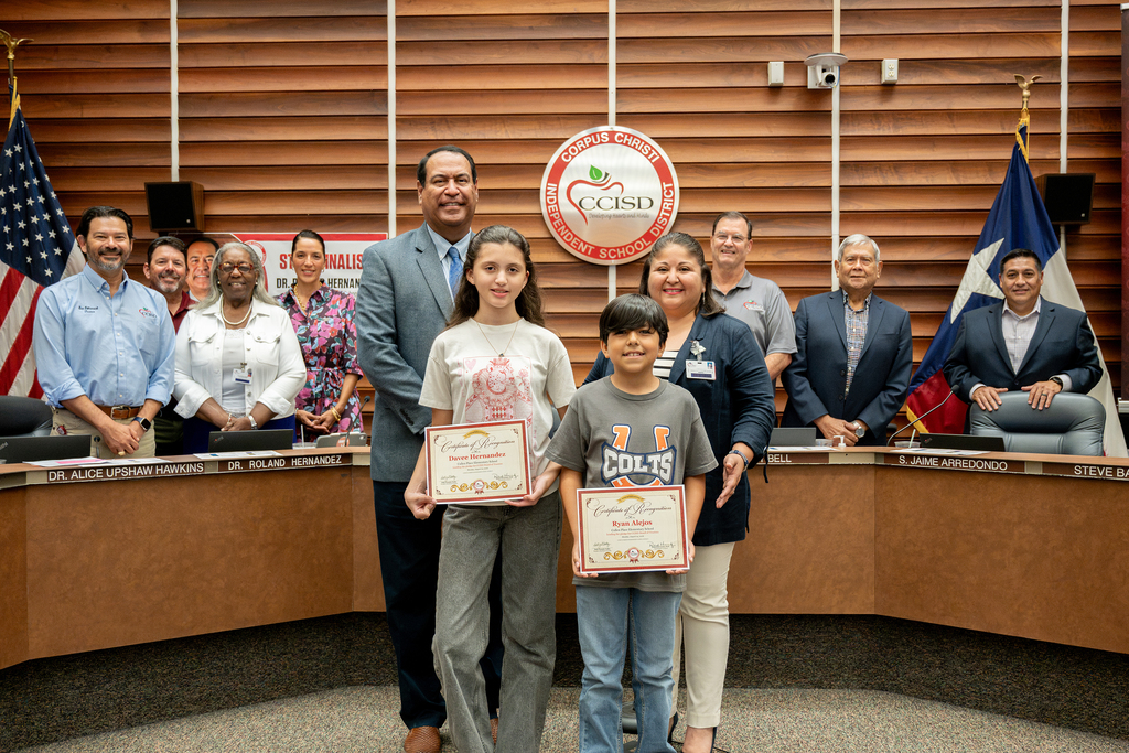 A group of adults and two children, one girl and one boy, hold certificates. They are standing in front of a desk.