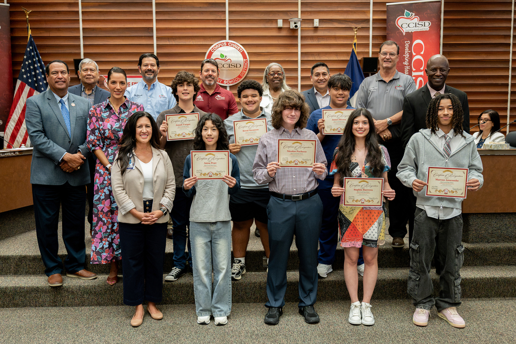 A group of students and officials stands on stairs, holding certificates. They wear casual attire. Behind them, three flags and a banner are displayed.