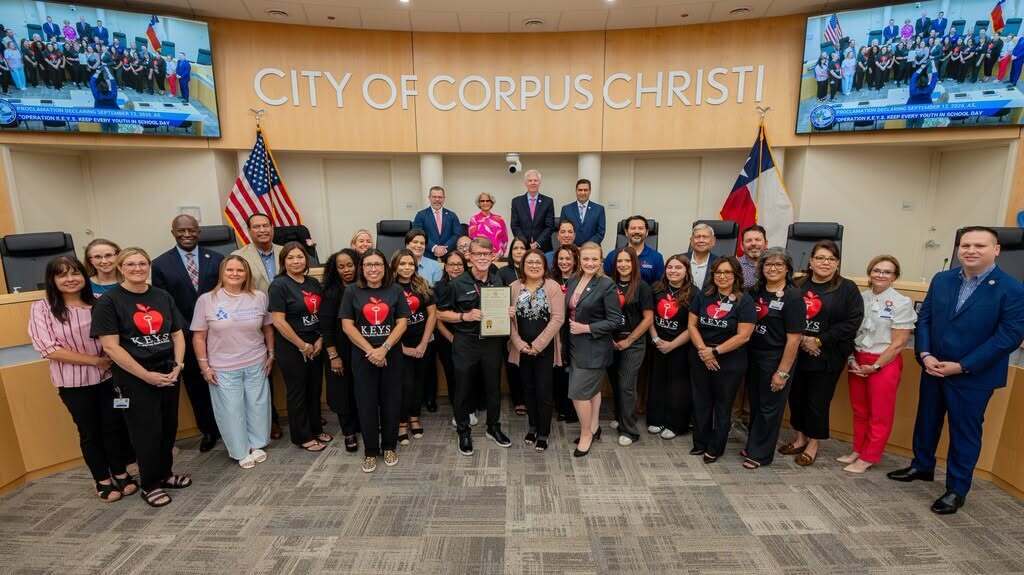 Group of people in suits, including officials, posing in a city council room with screens behind them.