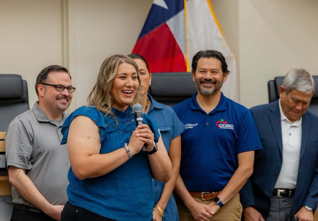 A group of adults stands in a room. A woman in a blue shirt speaks into a microphone.