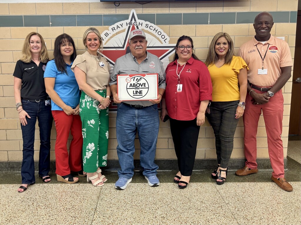 A group of adults stand behind a man holding a framed certificate, smiling. Behind them, a wall features a star and school name.
