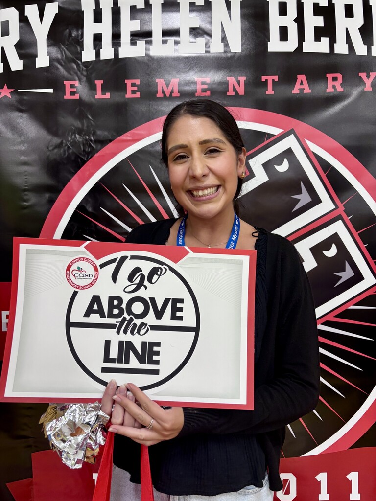 Woman holding a white sign with black text and a basketball graphic, smiling. Behind her is a banner with the text "RY HELEN BER ELEMENTARY".