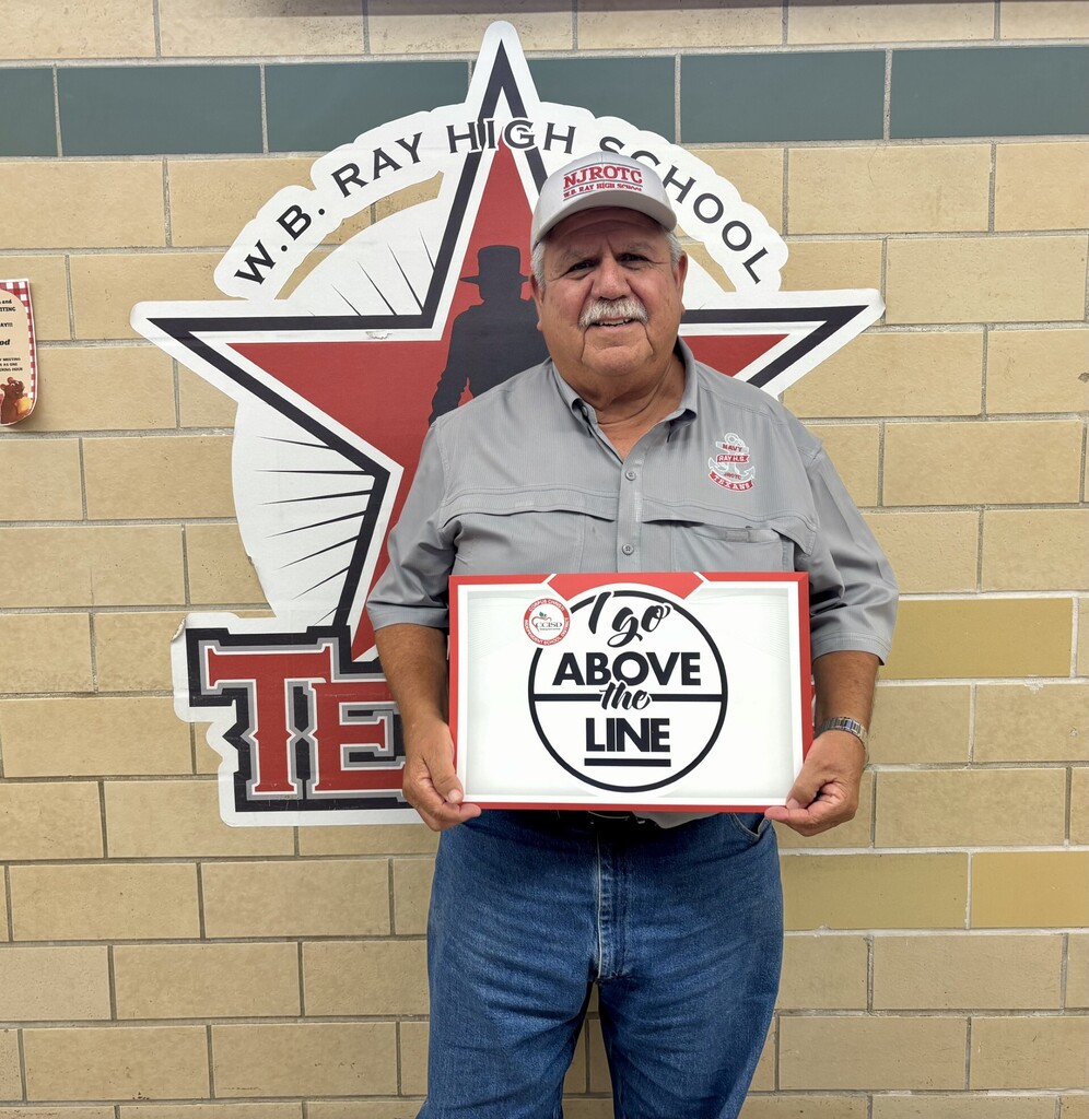 Man in a gray shirt and hat holds a sign that says "100 Above the Line" in front of a W.B. Ray High School sign.
