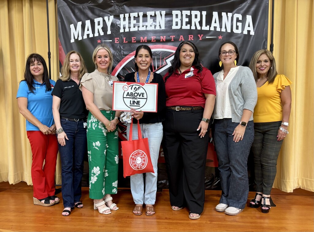 Seven women in different tops and pants, two holding a sign, standing together on a wooden floor in front of a banner.