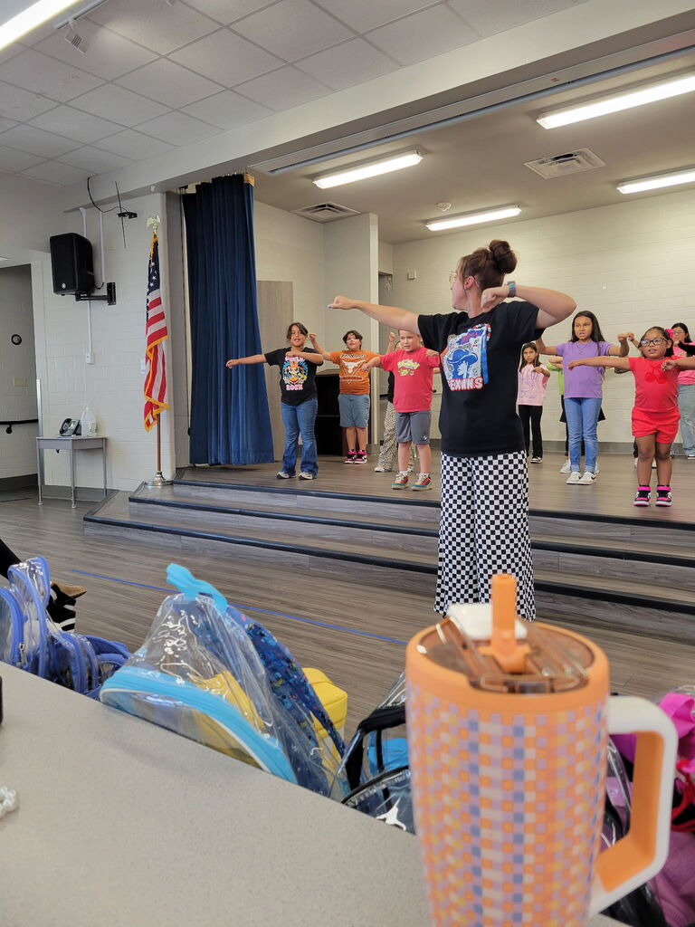 Group of children wearing shirts and sneakers performing dance moves with a woman in the front.