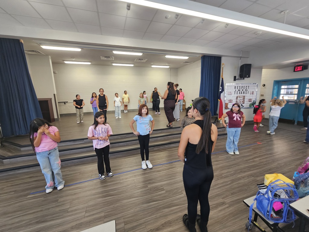 Children in a gym, some standing on a stage, with a woman in front, blue curtains, and a red sign.
