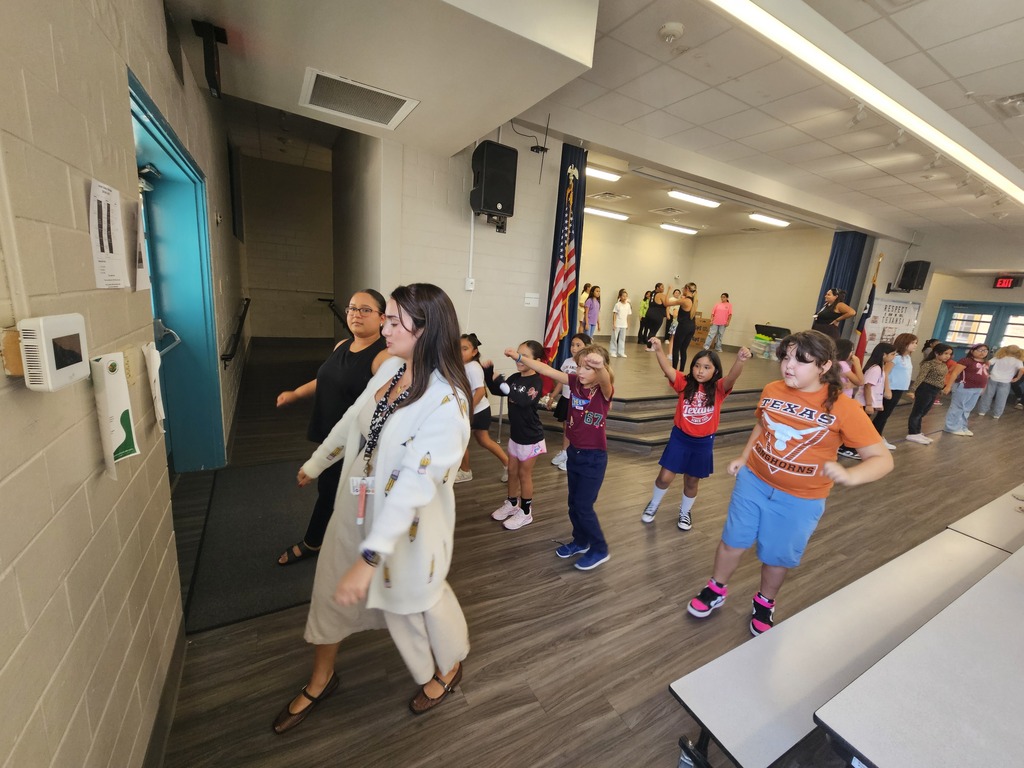 A group of children and two adults are dancing in a room with wooden floors, tables, and a flag.