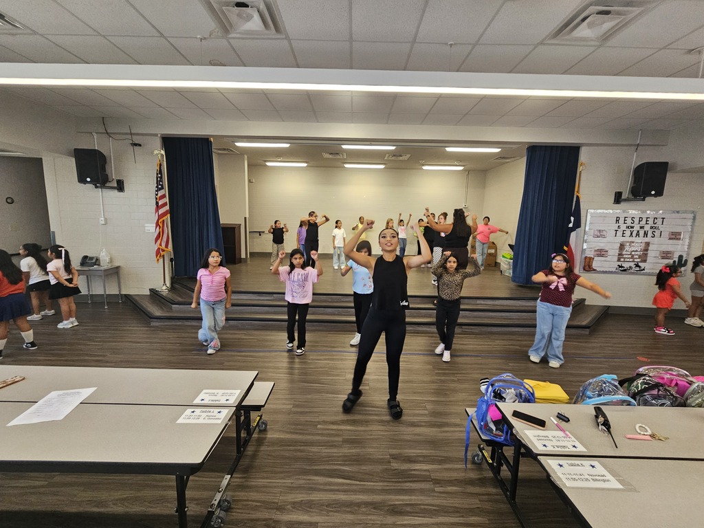 In a room with tables, children perform dance moves. A stage with curtains is behind them.