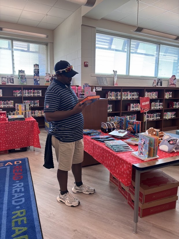 A man reads a book in a library. Shelves with books and a table with books are nearby.