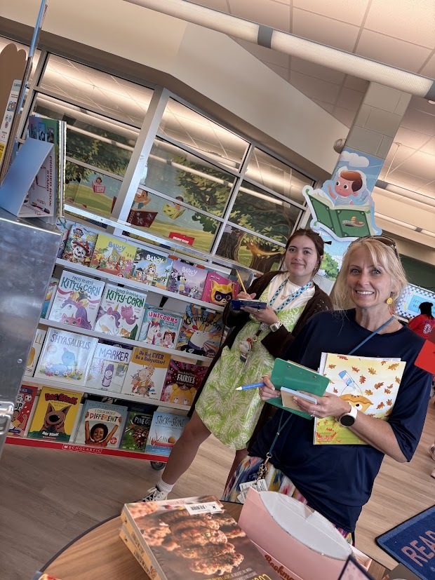 Two women in a bookstore, one holding a book and the other holding a book and pen. Books on shelves in background.