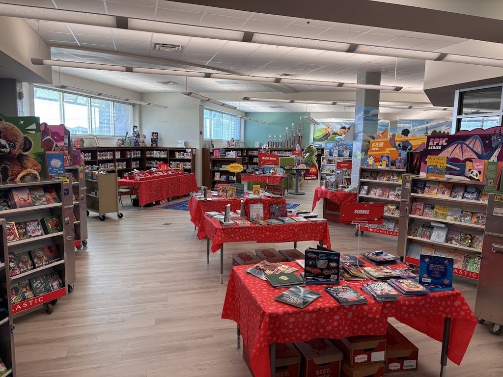 A library interior featuring tables covered with red cloth, books, and various items. Bookshelves line the walls, and overhead lighting illuminates the space.