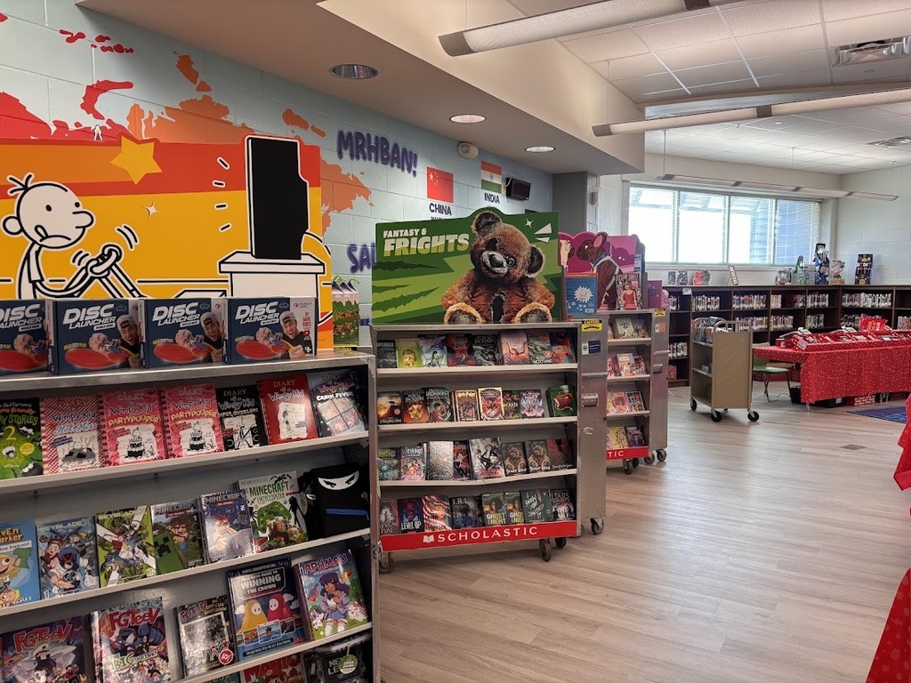 Interior of a library. Shelves with books, a wall with cartoon images, and ceiling lights. A table with a red tablecloth.