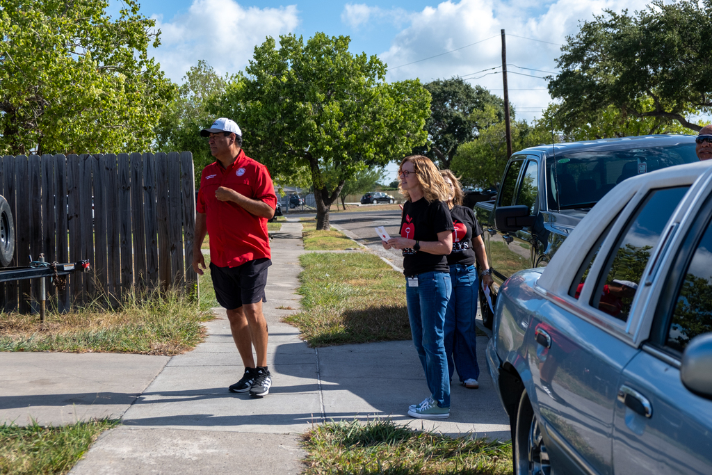 People in red and blue shirts stand on a sidewalk next to cars. A man walks towards them.