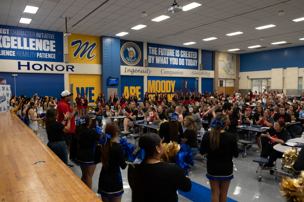 Large group of people in a room with cheerleaders performing. Bright lights, tables, and blue walls.
