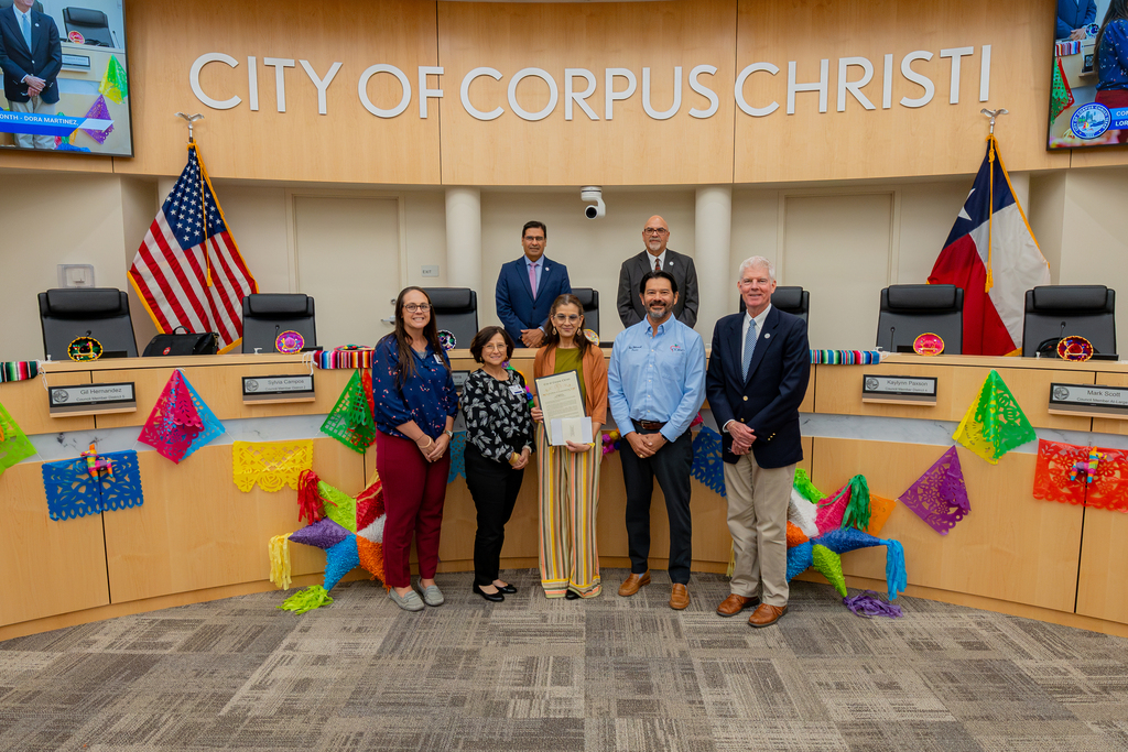 A group of people stand in a council chamber. Colorful flags hang behind them. A woman holds a certificate.