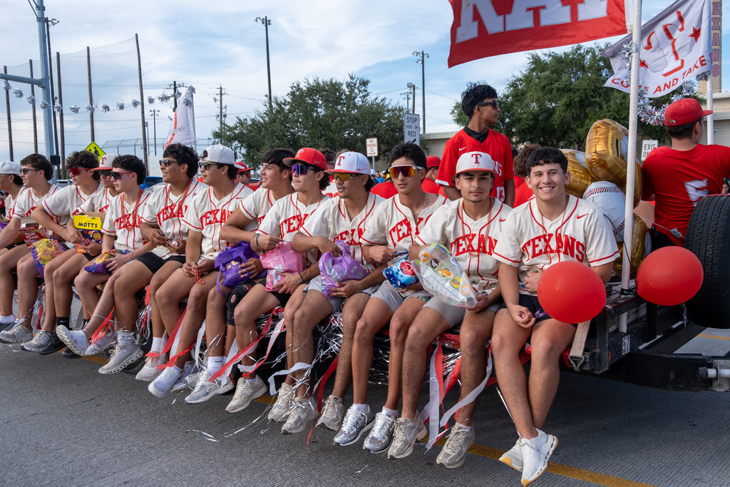 Athletes in white jerseys sit on a vehicle float, holding balloons and pom-poms, in a parade.