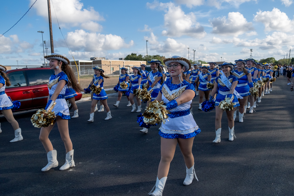 A group of cheerleaders in blue and white uniforms, holding pom-poms, walking in a parade on a road.