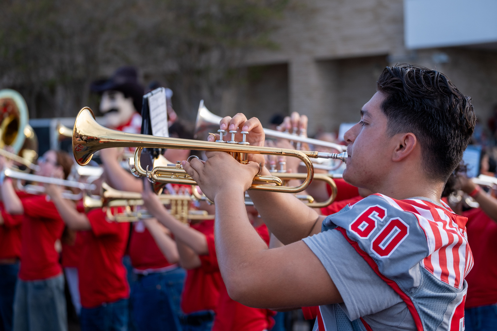 A person plays trumpet in a group, wearing a gray and red striped shirt with "60" on it.