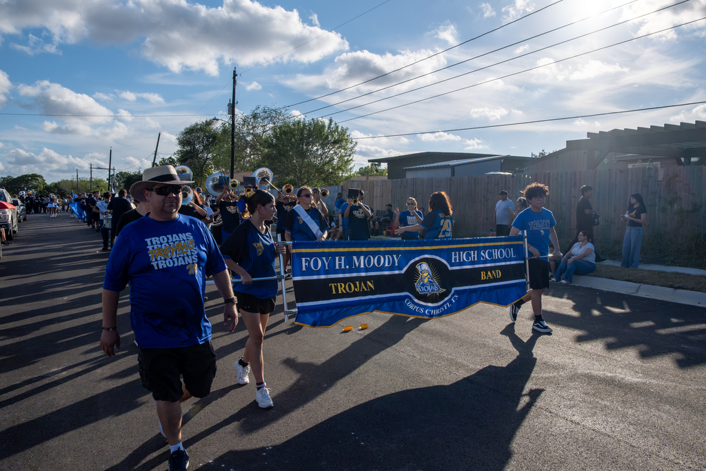 A marching band carrying a blue and white banner walks in a parade on a sunny day.