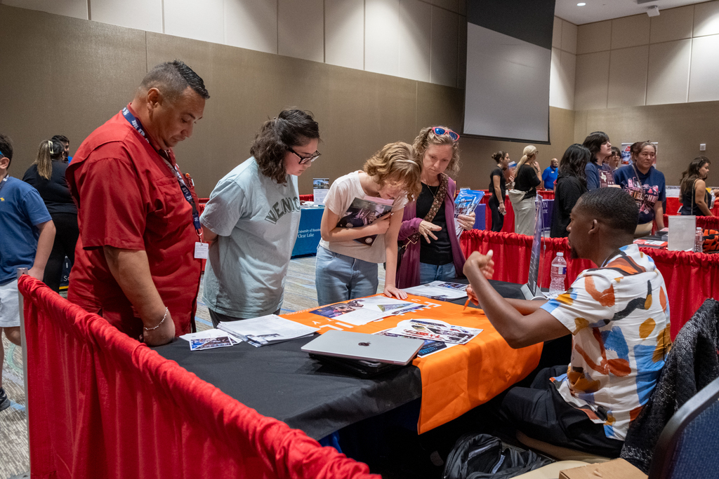 People gather at a table with a red and black cloth, looking at papers and a laptop.
