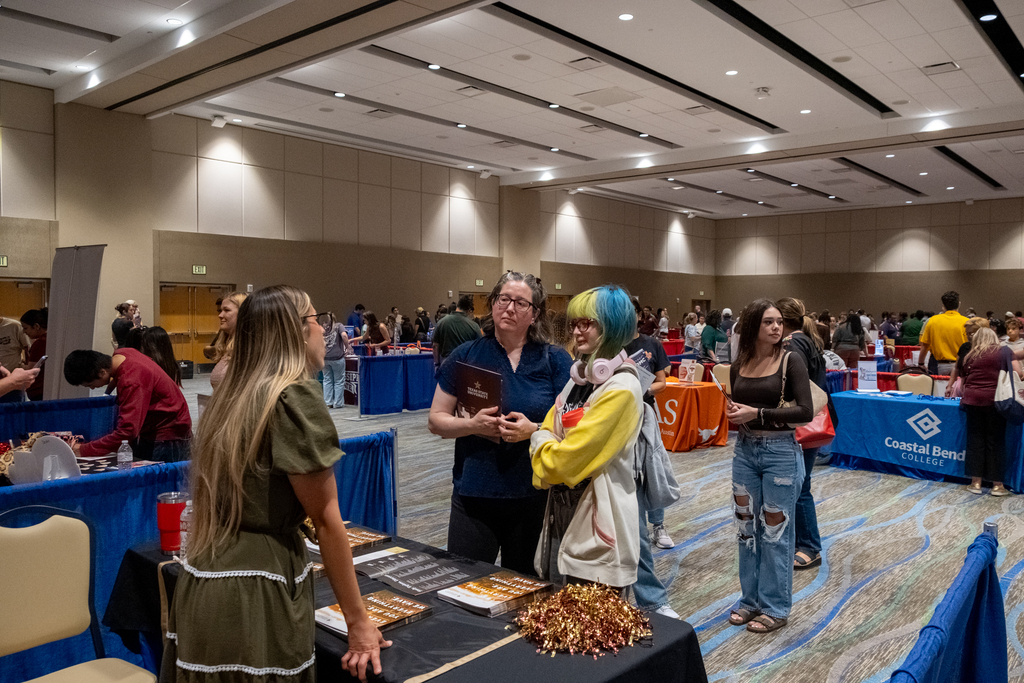 A large room with tables and people at a career fair. A woman in colorful hair speaks with others.