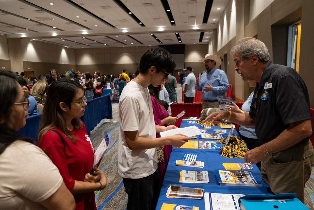 Multiple people stand around a table with brochures and pamphlets. A man wears a badge.