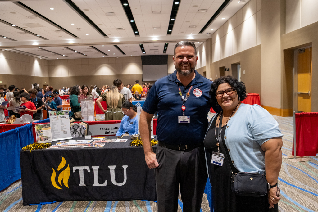 A man and woman stand together in a conference hall, smiling. The table before them displays a TLU logo.