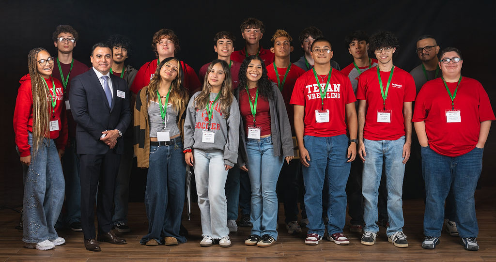 A group of students and adults stand on a wooden floor. They wear red shirts with green lanyards.