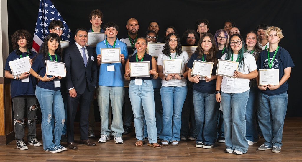 A group of people, some holding certificates, pose for a picture on a stage with an American flag.