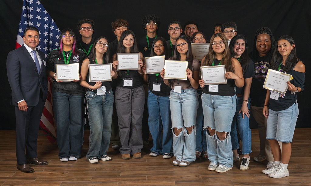 A group of students and a man holding certificates stand together, posing for a photo in front of a backdrop with an American flag.