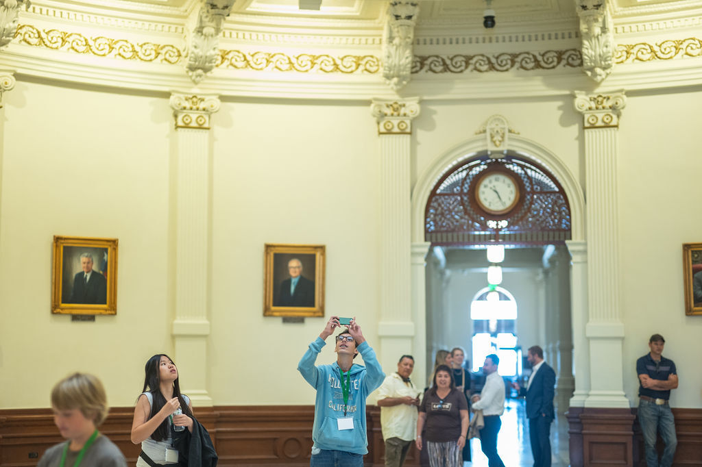 People in a room with arched doorways, wooden paneling, and framed portraits. One person takes a photo.