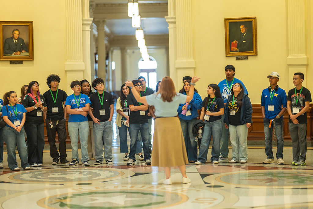 A woman leads a group of young people in a large room with decorative columns and a patterned floor.